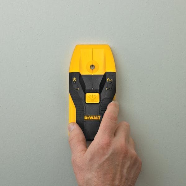 A hand holding a yellow and black DEWALT electronic stud finder against a light gray wall.