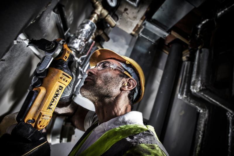 Worker uses a DEWALT DWE315 tool to cut wall pipe, wearing safety helmet and vest in industrial setting.