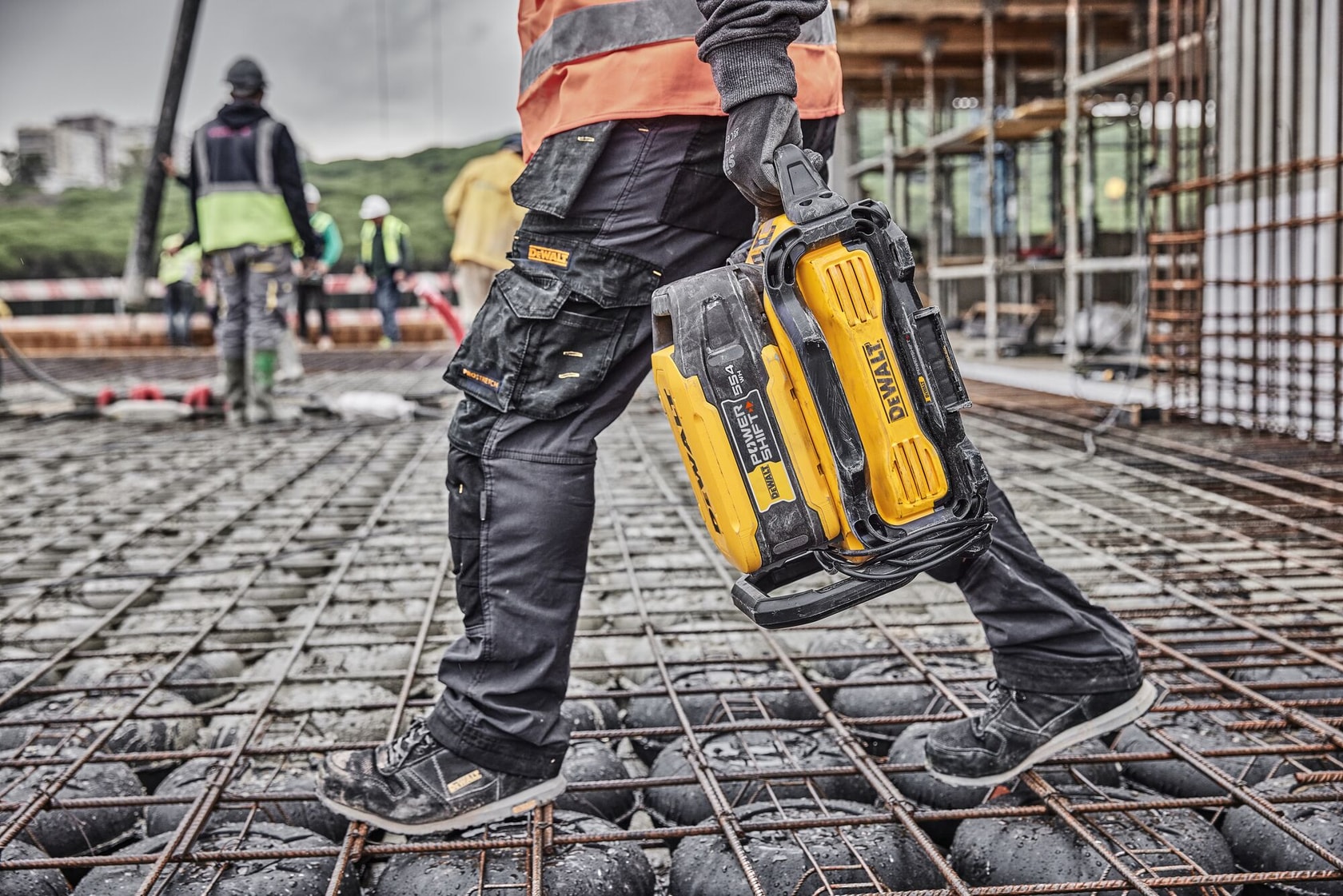 Worker carries a yellow DEWALT battery on a construction site with steel grid flooring.